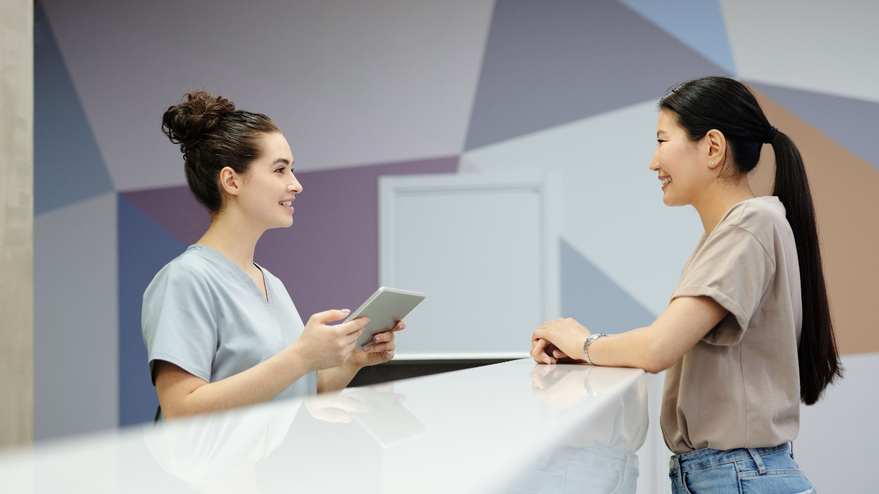 A clinic receptionist is speaking with a patient across the front desk. Photo by Cedric Fauntleroy, sourced from Pexels.com