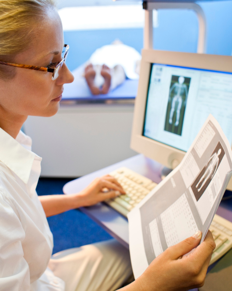 A technologist examines a bone density scan printout while a patient lies on an exam table in the background. Photo by Science Photo Library, sourced from Canva