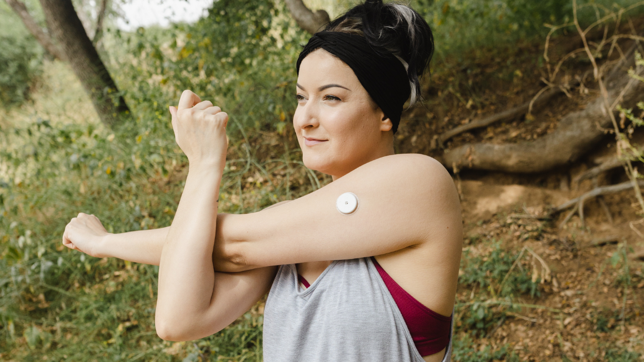 A woman stretching before a run, wearing a continuous glucose monitoring device on her upper arm. Photo by Maya Holt with baseimage, sourced from Canva.
